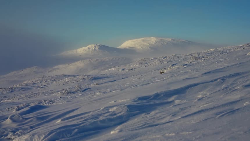 Strong winds create a ground blizzard, blowing snow across a vast, frozen mountain landscape under a clear sky