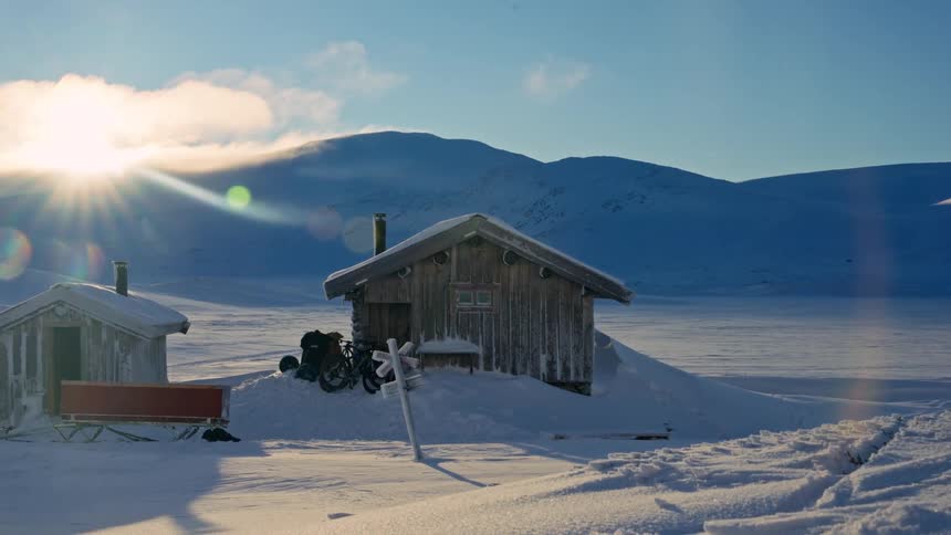 A remote wooden cabin sits nestled in deep snow in the arctic wilderness during a bright, sunny winter day
