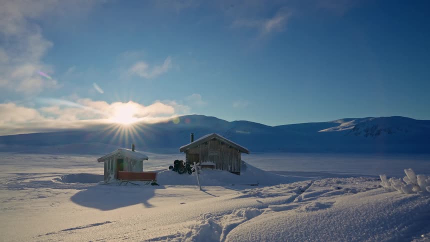 Remote wooden cabins are bathed in the warm light of the rising sun in a vast, snowy, and frozen landscape