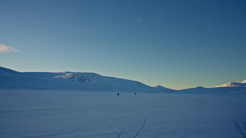Two cross-country skiers traverse a vast, frozen landscape with snow-covered mountains in the distance under a clear blue sky