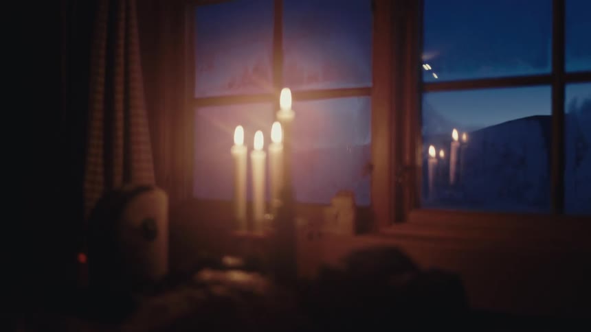 A cozy winter evening scene with flickering candles on a windowsill casting a warm glow on a frosty window as the view softly blurs