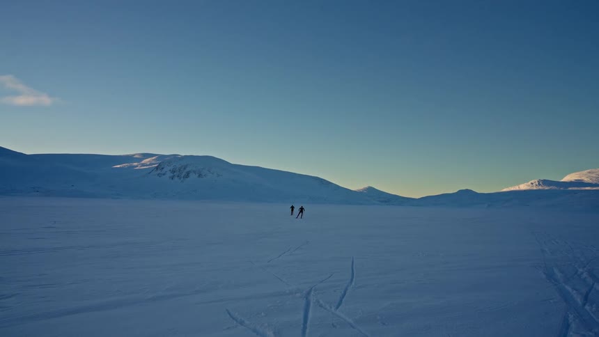 Two distant cross-country skiers traverse a vast, flat, snowy landscape towards mountains under a clear blue sky