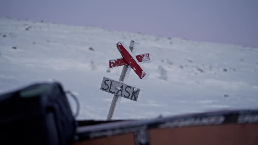 A frozen wooden trail marker sign covered in ice stands in a vast, desolate, snow-covered landscape