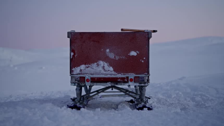 A static shot of a red snow sled trailer parked in a desolate, snowy landscape under a winter sky