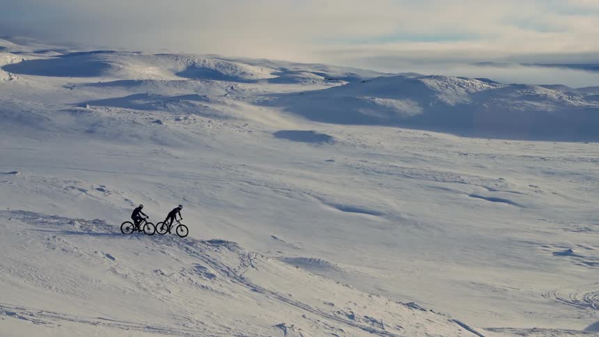 Two silhouetted cyclists ride their fat bikes across a vast, snowy, and rolling landscape before stopping to take in the view
