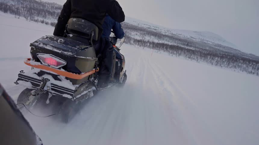 A first-person perspective shows a snowmobile following another through a vast, snow-covered arctic landscape