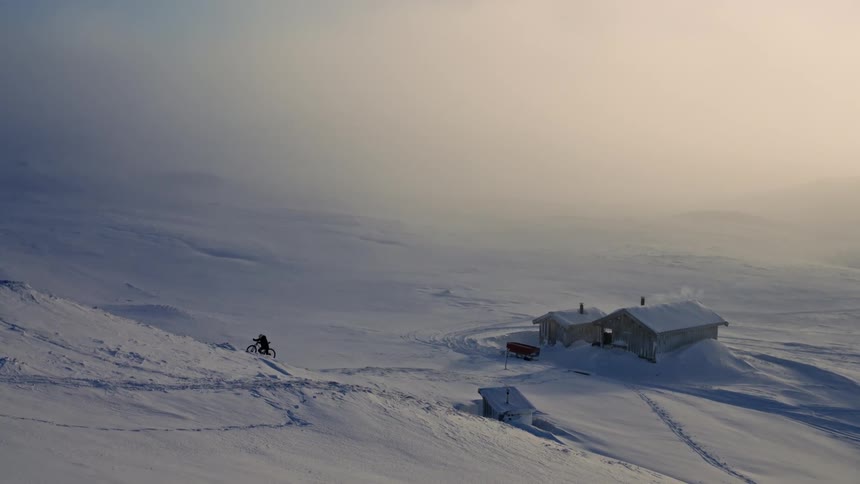 A person pushes a fat bike up a vast, snowy slope towards a remote cabin in a foggy winter landscape