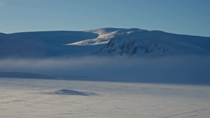 Sunlight illuminates snow-covered mountains behind a misty, frozen valley where wind blows snow across the arctic tundra