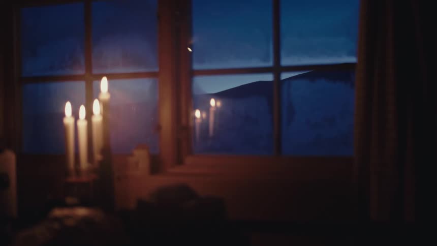 A soft-focus shot captures the cozy glow of flickering candles by a window, with a snowy winter scene visible outside at dusk