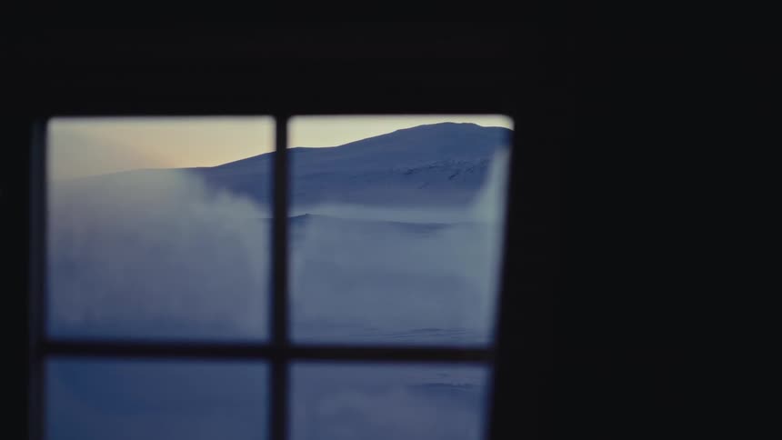 A view from a window inside a dark room shows a snow-covered mountain landscape with strong winds blowing snow across the ground