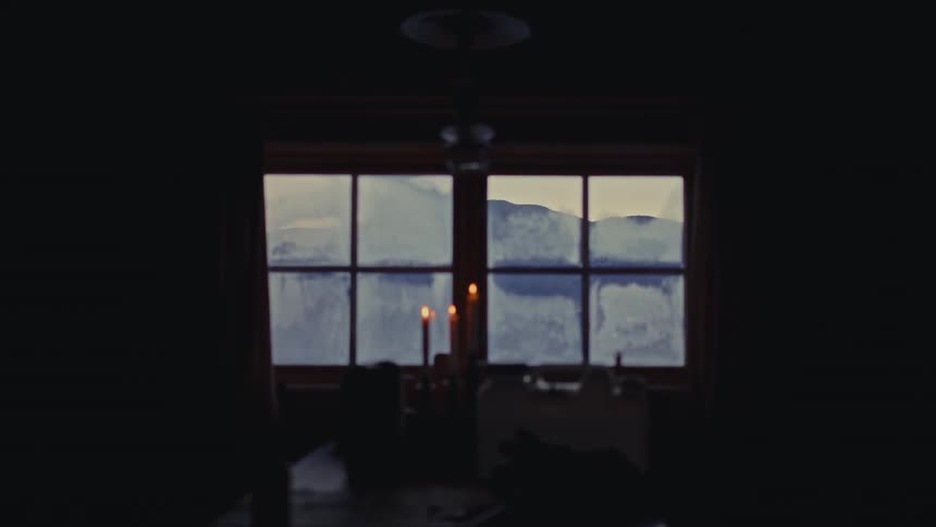 A soft-focus view from a dark room shows lit candles flickering in front of a frosted window looking out at a snowy mountain landscape