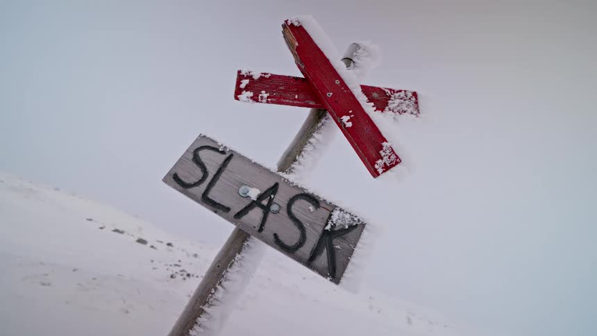 A frozen wooden signpost with a red trail marker and the word 'SLASK' stands alone in a desolate, snow-covered mountain landscape