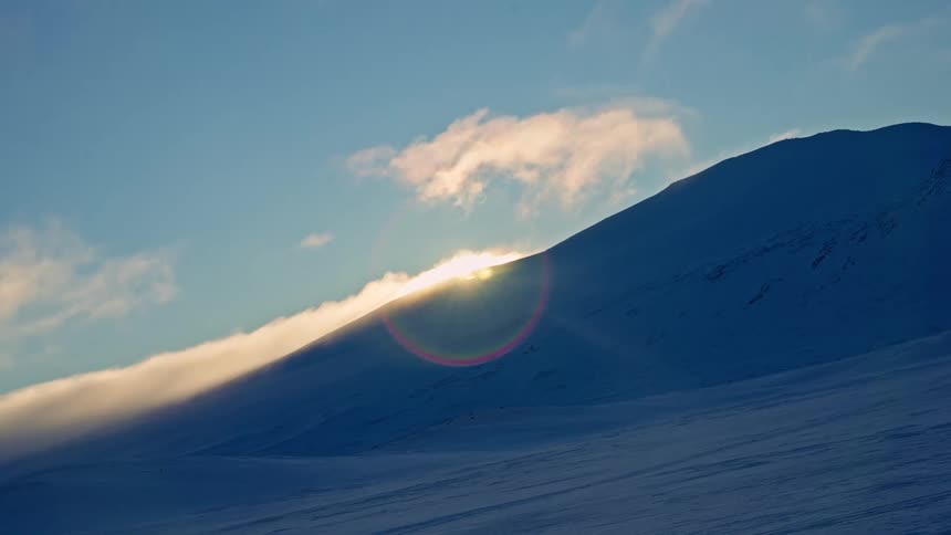 A breathtaking sunrise crests over a vast, snowy mountain landscape as clouds drift gently past