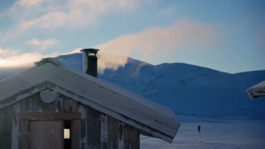 A cozy, snow-covered cabin with smoke rising from the chimney stands in a vast, frozen mountain landscape as a lone cross-country skier glides by in the distance