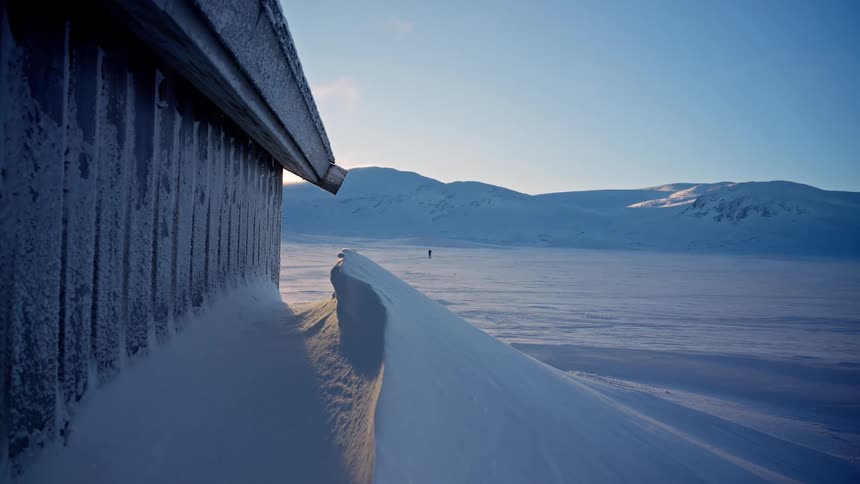 A lone cross-country skier glides across a vast, snowy mountain landscape next to a frost-covered wooden cabin in the cold morning light