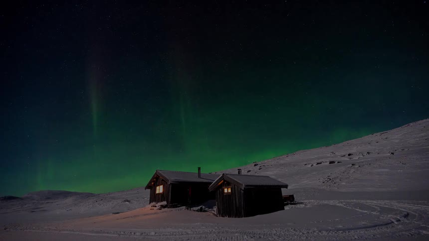 A stunning timelapse shows the green Aurora Borealis dancing in the starry night sky above two cozy, snow-covered cabins in a remote winter landscape