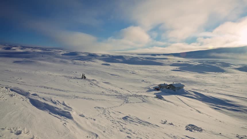 An expansive, sunlit snowy landscape with a remote cabin and two people with skis standing still