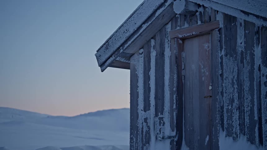 A close-up of a weathered wooden cabin covered in frost and snow, standing alone in a vast winter landscape at twilight