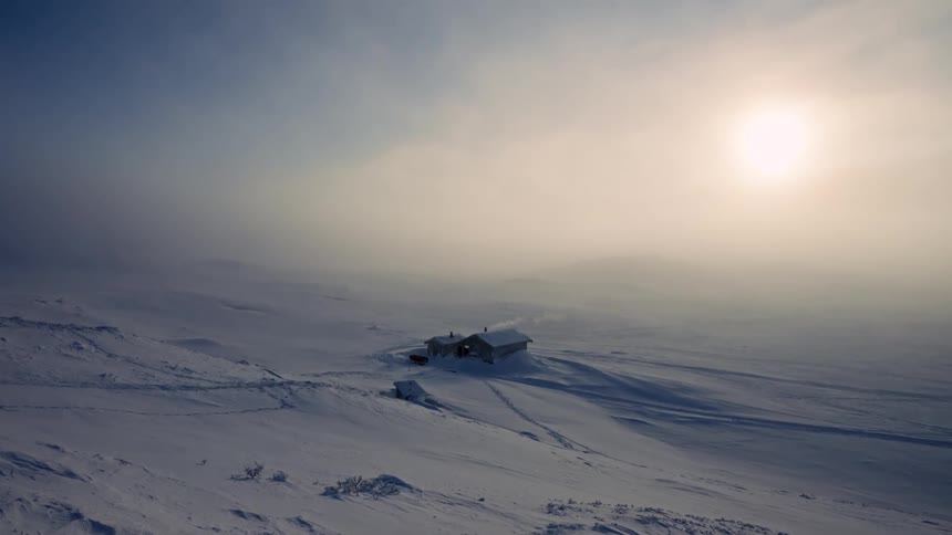 A secluded cabin sits isolated in a vast, snow-covered landscape as the sun glows through the thick winter fog
