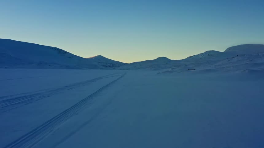Aerial footage pulls back over tracks in a vast, snow-covered mountain valley during the serene blue hour