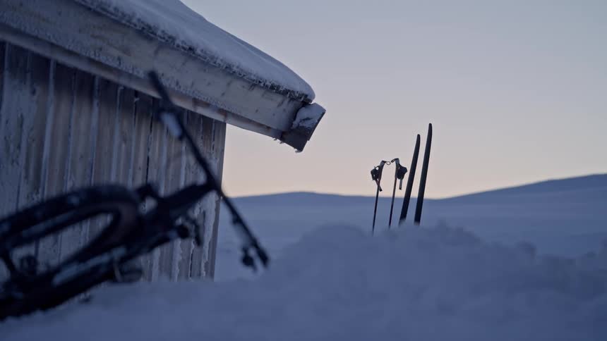 A cinematic rack focus from a fat bike to a pair of skis resting in deep snow outside a rustic cabin at dusk