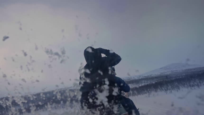 A person in a hooded jacket rides a snowmobile through a vast, snowy landscape, filmed from a low angle on the vehicle with snow obscuring the lens