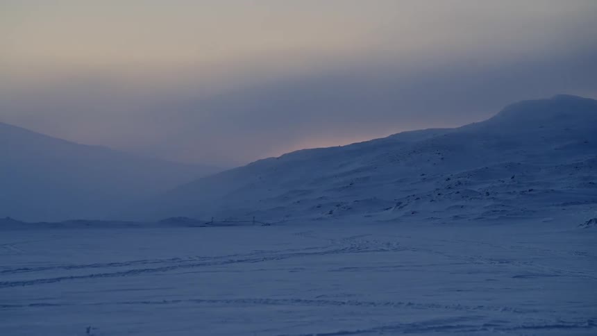 A serene panning shot reveals a vast, snow-covered mountain landscape during a cold and misty twilight