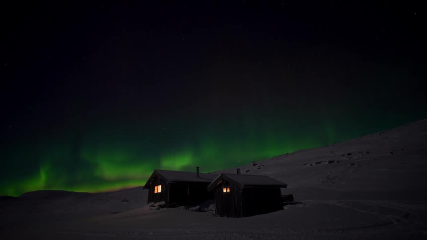 A stunning timelapse of the green Northern Lights dancing in the starry night sky over two cozy cabins in a remote, snowy landscape