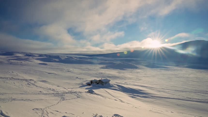 The sun shines brightly over an isolated cabin nestled in a vast, desolate, and snow-covered landscape