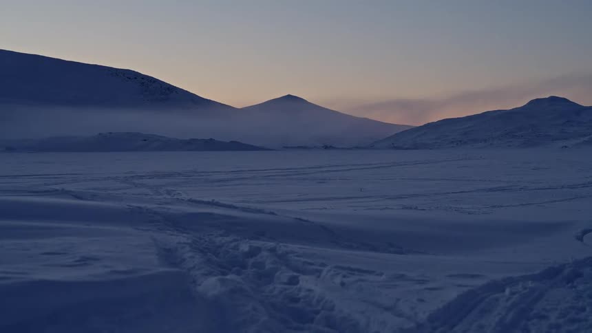 A slow pull-back shot reveals a vast, frozen arctic valley and distant misty mountains during a serene twilight