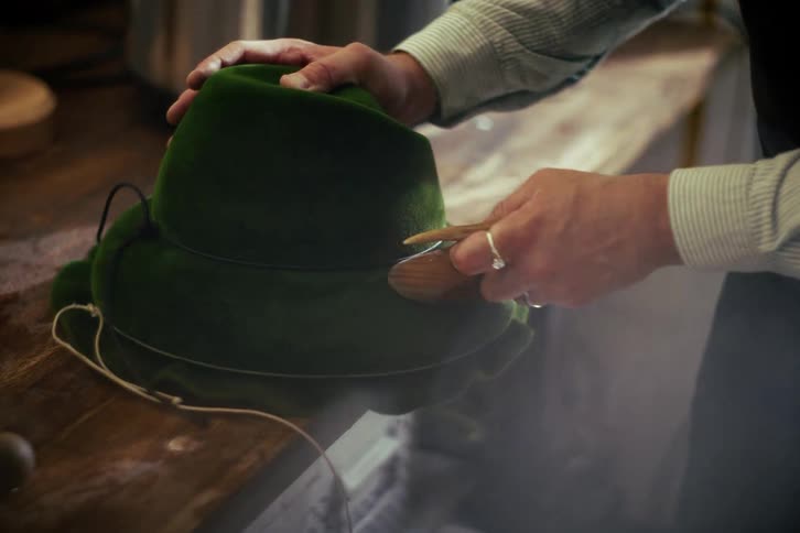 A skilled artisan meticulously brushes and steams a traditional green felt hat in their workshop