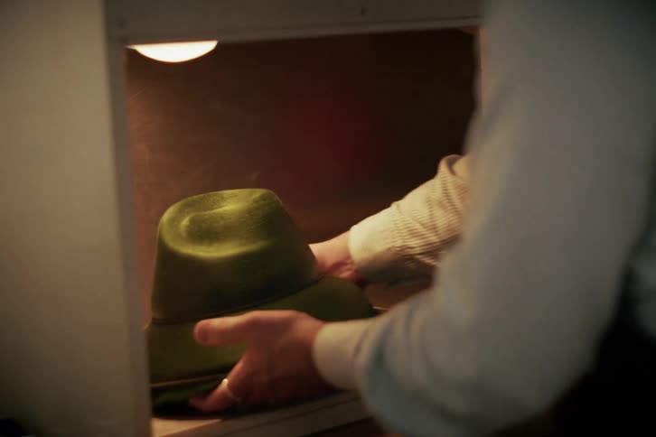 A hatter opens a lit cabinet and carefully places a newly finished green felt hat inside for setting