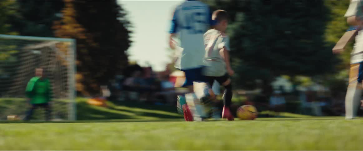 A dramatic low-angle slow-motion shot captures the intense action of a children's soccer game on a sunny day