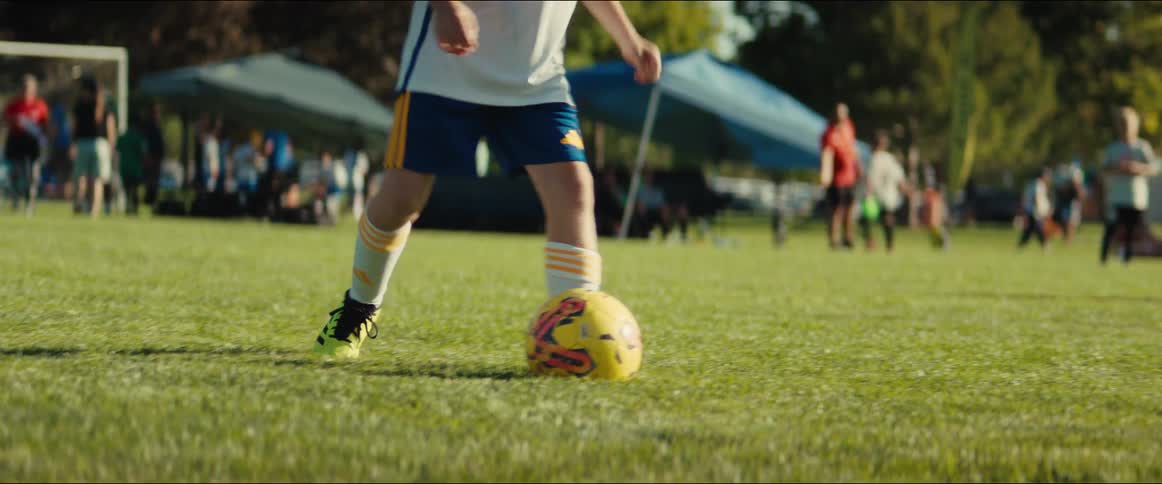 Low angle shot of a child's legs kicking a soccer ball on a green field during a youth league game