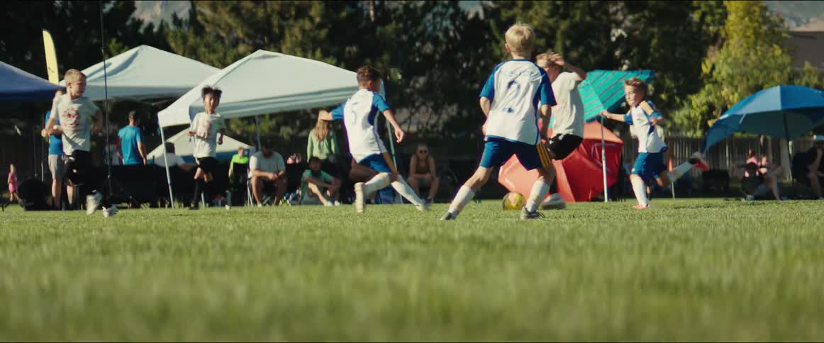 A low-angle, slow-motion shot of a children's soccer game with parents watching from the sidelines on a sunny day