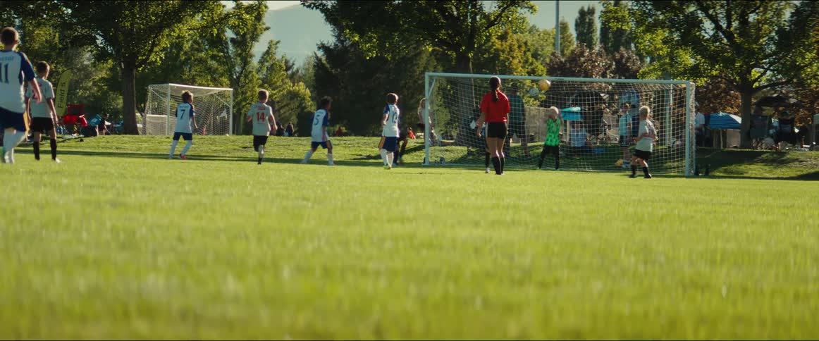 In slow motion, a young soccer player kicks the ball toward the goal as the goalie makes a save during a youth league game on a sunny day