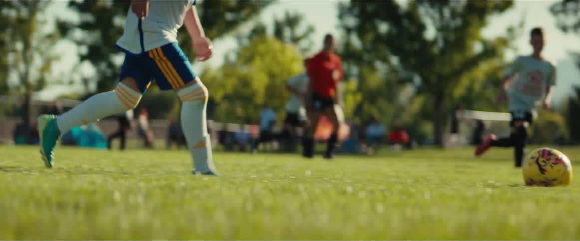 In slow motion, a young goalie tosses the ball into play and a teammate dribbles and kicks it down the field during a sunny kids' soccer game