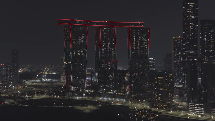A striking modern building with three towers connected by a skybridge glows with red lights against a dark night cityscape