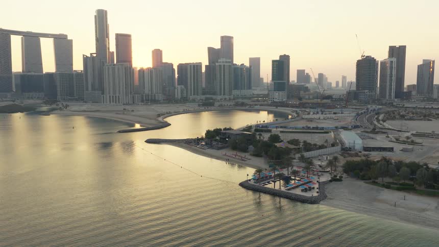 An aerial view shows the sun rising behind the modern skyscrapers of Abu Dhabi's skyline, with calm waters and a beach club in the foreground