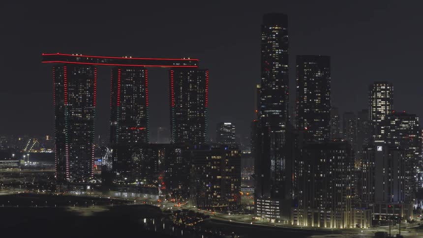 An aerial view of the illuminated Abu Dhabi city skyline at night, featuring the iconic red-lit Gate Towers as the camera slowly zooms out