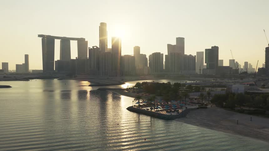 A serene aerial shot captures the golden sunrise over the Abu Dhabi city skyline, reflecting on the calm waters next to a peaceful beach resort