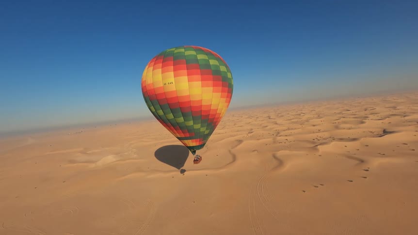 A vibrant, multi-colored hot air balloon glides gracefully just above the golden sand dunes of a vast desert under a clear blue sky