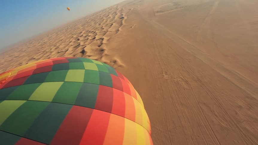 POV shot from the top of a vibrant hot air balloon as it glides down towards the expansive desert dunes below