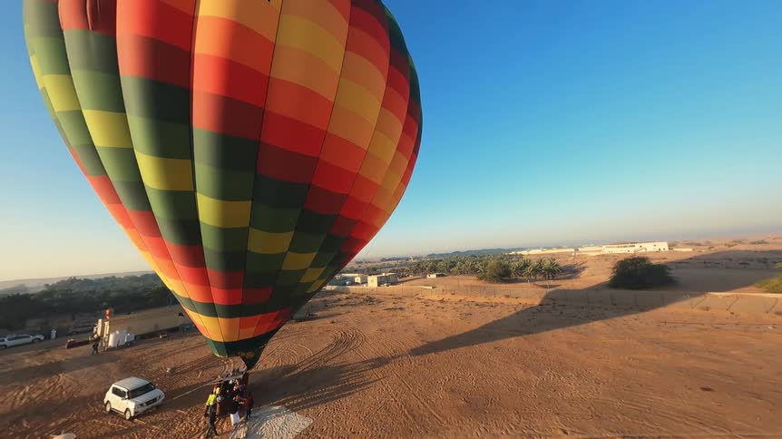 A colorful hot air balloon slowly ascends from the desert ground as people watch from below