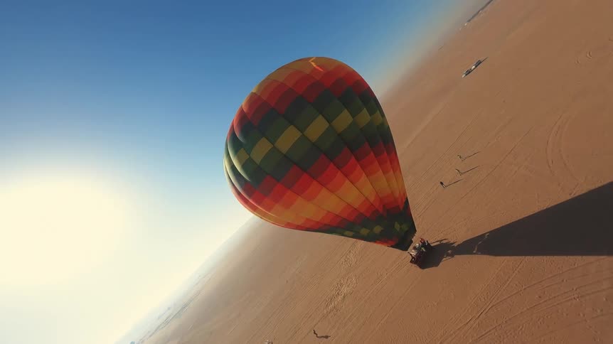 An immersive aerial view captures a colorful hot air balloon taking off and rising above the vast desert landscape under a brilliant sun
