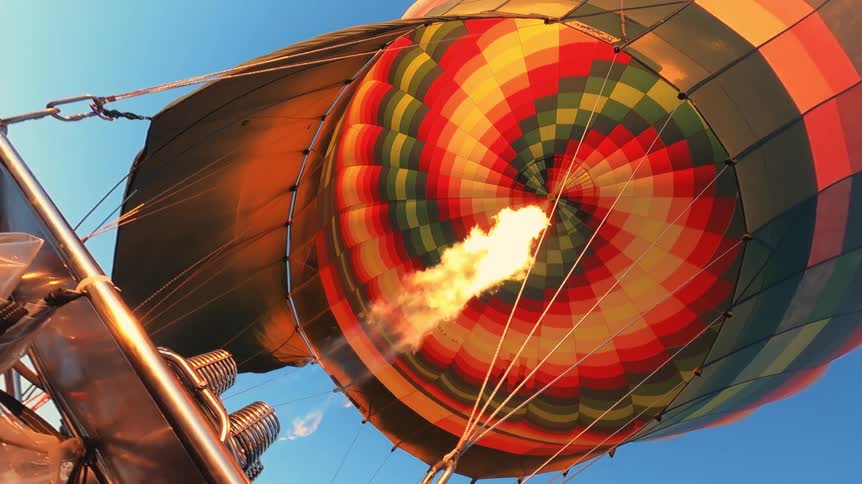 A close-up, slow-motion view from the basket shows a powerful flame from the burner inflating a colorful hot air balloon against the clear blue sky