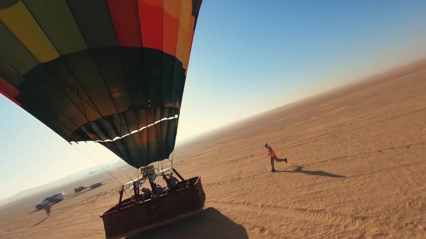 An aerial view shows a colorful hot air balloon with passengers lifting off and ascending over a vast sandy desert on a sunny day