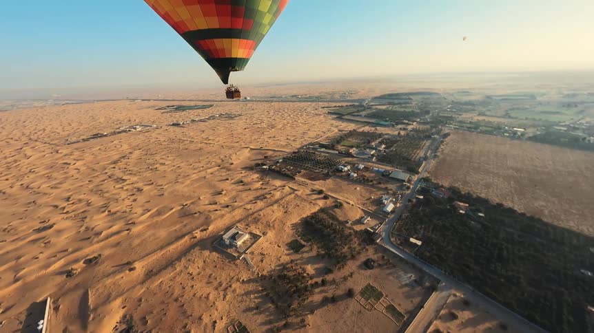 A colorful hot air balloon full of tourists flies over the vast golden desert sand dunes on a clear sunny day