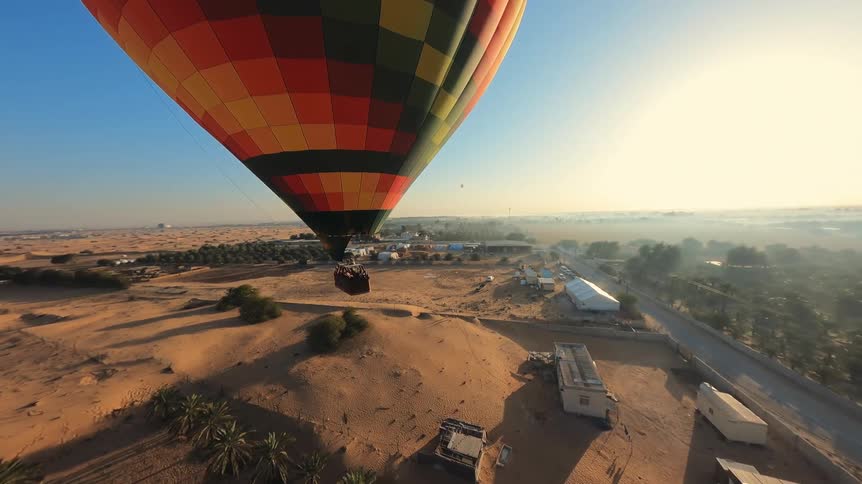 An FPV drone captures a stunning aerial view of a colorful hot air balloon gracefully flying over a desert oasis at sunrise