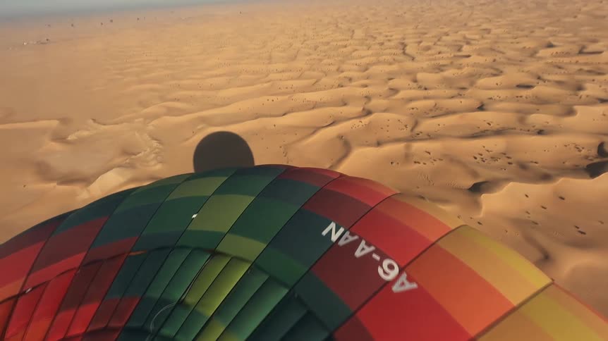 Soaring high in a colorful hot air balloon, this aerial footage captures the stunning expanse of desert dunes under a bright, clear sky